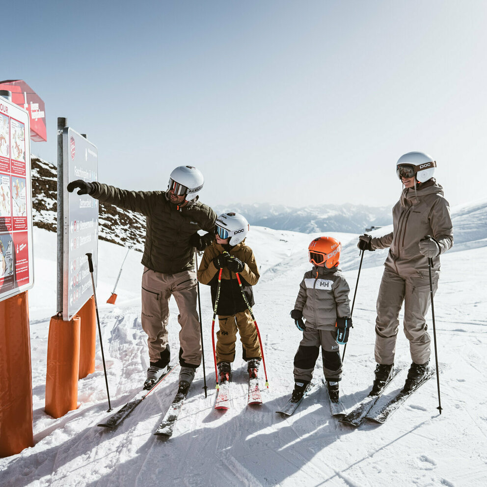Familie mit zwei Kindern auf Skiern vor einer Informationstafel im Skigebiet Zettersfeld in Osttirol. Der Vater zeigt auf die Sicherheitshinweise, während die Familie aufmerksam zuhört.