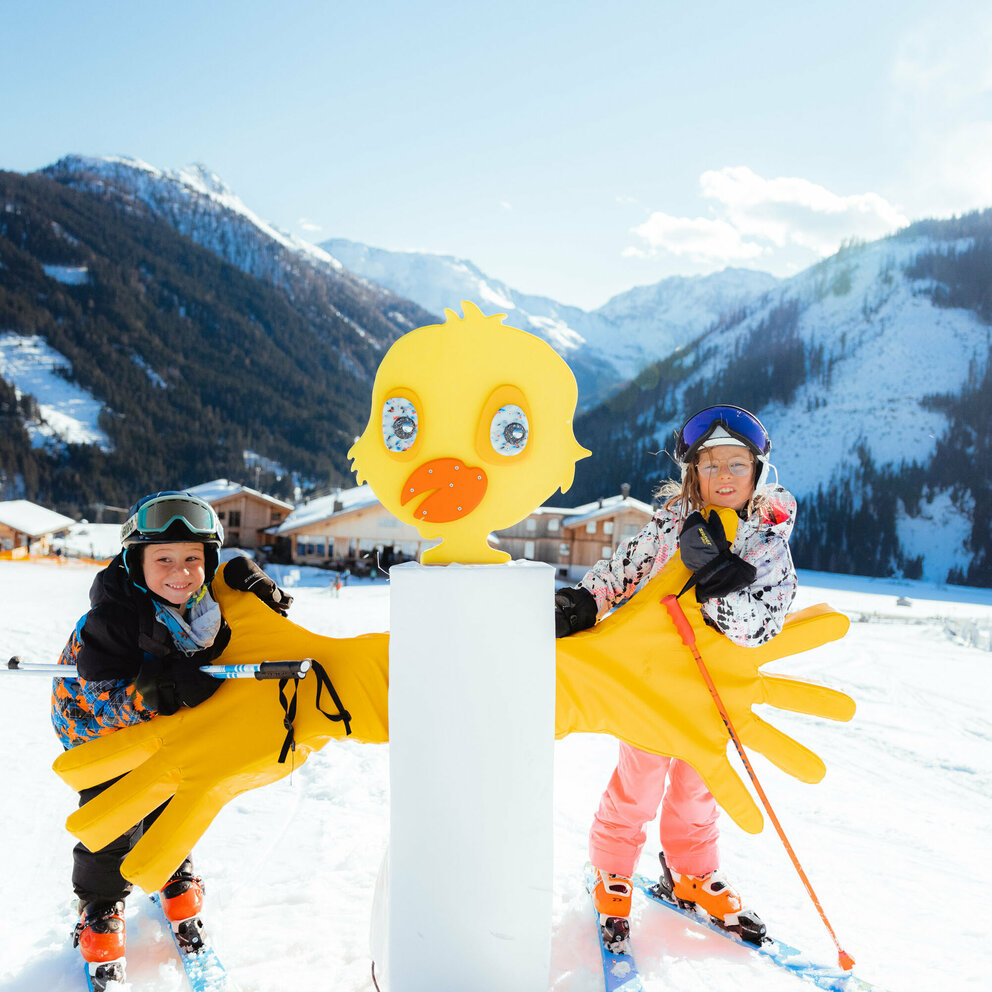 Zwei Kinder stehen im familienfreundlichen Skigebiet Golzentipp in Obertilliach hinter einem gelben Kurvenzeiger in Entendesign mit winterlichem Bergpanorama im Hintergrund.