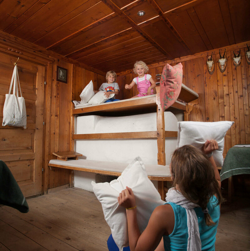 Eine Frau sitzt auf dem Boden in der Stube und hält zwei Kissen in der Hand. Auf dem Ofen sitzen zwei Kinder und werfen Kissen auf die Frau.