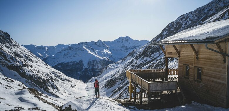 Ein Skitourengeher steht neben der hölzernen Eisseehütte mit Blick talauswärts. Sie Sonne strahlt am blauen Himmel.