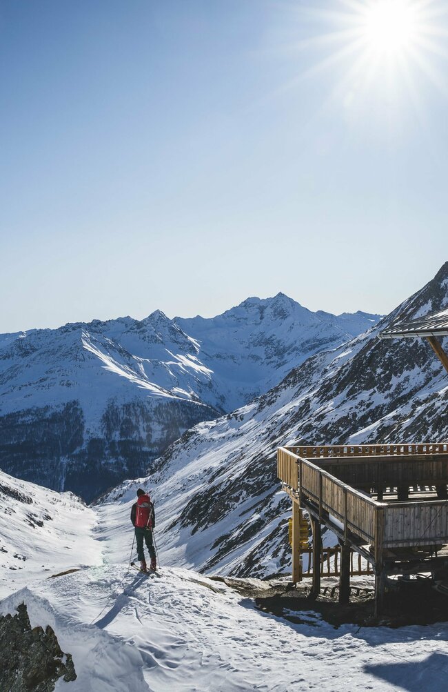 Ein Skitourengeher steht neben der hölzernen Eisseehütte mit Blick talauswärts. Sie Sonne strahlt am blauen Himmel.