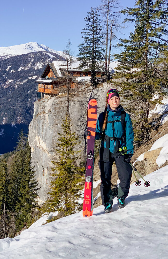 Alpine Sicherheit Eine Skitourengeherin steht im Schnee und hält ihre Ski in den Händen, mit der Dolomitenhütte im Hintergrund.
