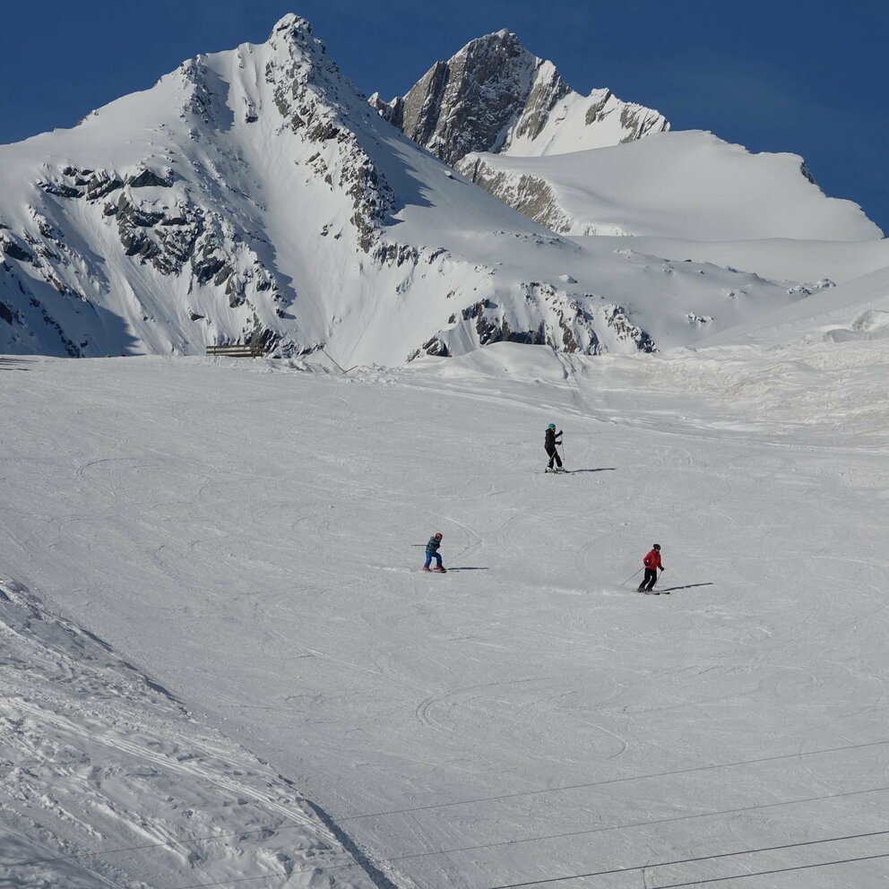 Mehrere Skifahrer auf einer breiten Piste im Skigebiet Bichllift in Prägraten. Im Hintergrund ragen zwei schneebedeckte Gipfel in den blauen Himmel.