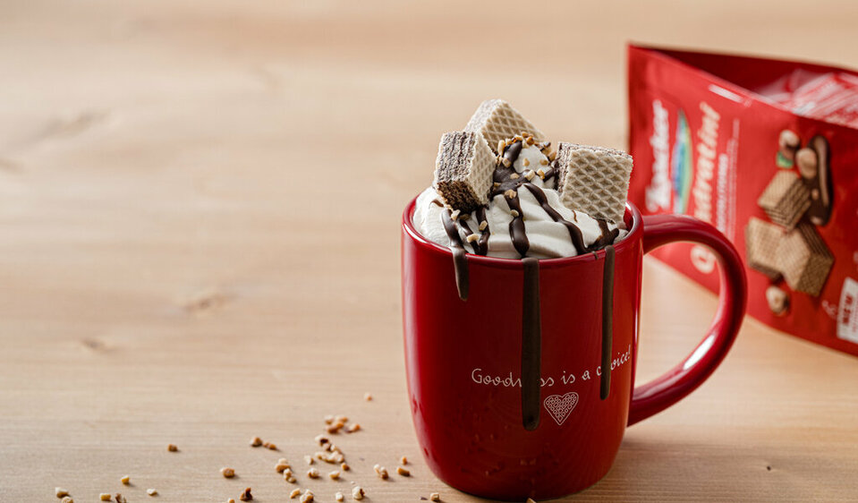 Die verführerisch große, rote Kakaotasse mit Sahnehaube und Schokosauce wird von drei kleinen Waffeln gekrönt, während im Hintergrund die bekannte rote Waffelverpackung von Loacker hervorlugt.