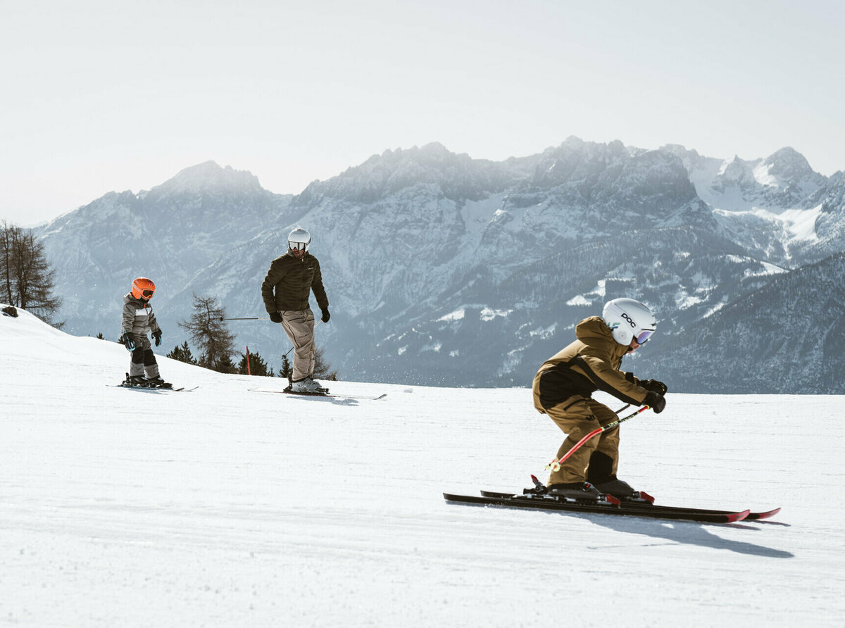 Vater mit zwei Kindern beim Skifahren auf einer verschneiten Piste im Skigebiet Zettersfeld in Osttirol, mit Blick auf die beeindruckende Bergkulisse der Lienzer Dolomiten.