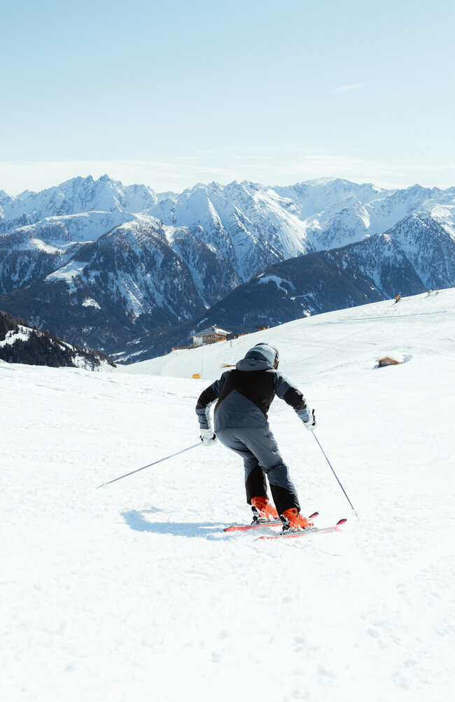 Zwei Skifahrerinnen auf einer flachen Piste oberhalb der Baumgrenze. Im Hintergrund der Taleinschnitt und ein verschneiter Bergkamm, der den Abschluss zur milchig wirkenden Bewölkung an einem Schönwettertag im Winter zeigt.