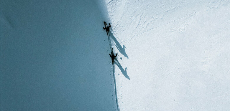 Unterwegs im Schnee, zwischen oben und unten, zwischen Licht und Schatten. Zwei Skitourengeher gehen auf ihren Skiern entlang einer Schattenlinie, die der Berg auf die frisch verschneite Landschaft wirft.