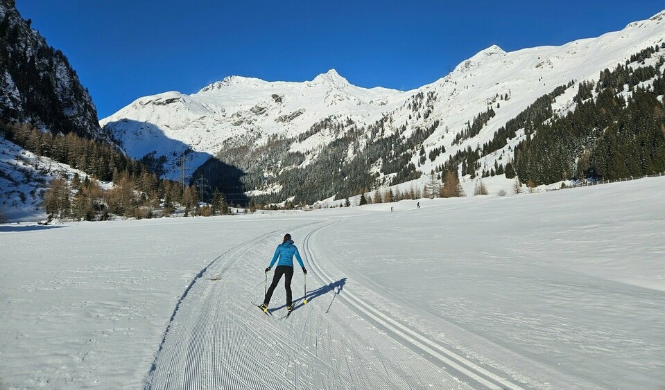 Eine Langläuferin fährt auf einer präparierten Loipe durch ein sonniges, schneebedecktes Tal, umgeben von hohen weißen Bergen und einzelnen Waldhängen. Weitere Langläufer sind in der Ferne zu sehen.