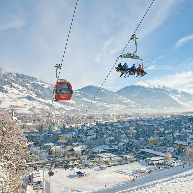 Sessel und rote Gondel schweben über die tief verschneite Winterlandschaft am Hochstein mit der Stadt Lienz im Hintergrund.