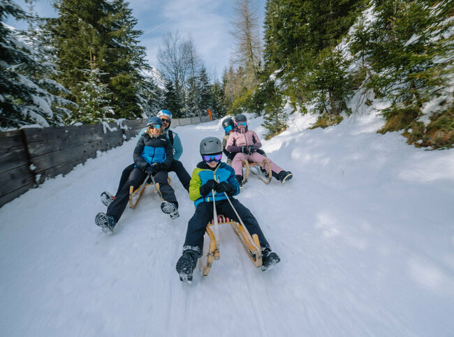 Fünf Personen in Skianzügen rodeln auf ihren Schlitten talwärts auf der Rodelbahn im Winkeltal in Außervillgraten, Osttirol.