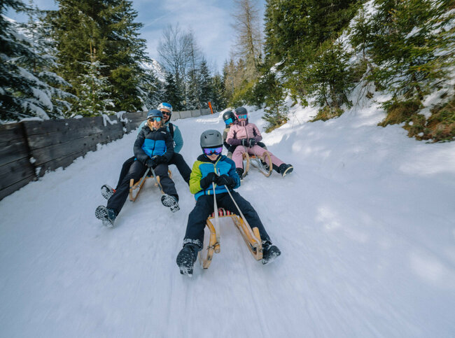 Eine Familie in Skianzügen rodelt auf ihren Schlitten talwärts auf der Rodelbahn im Winkeltal in Außervillgraten, Osttirol.