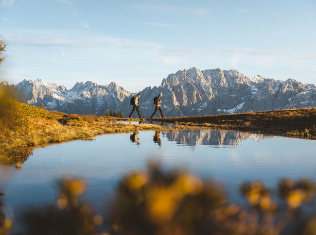 Der See am Hochstein spiegelt zwei vorbei Wandernde und die Bergkulisse in seinem klaren Wasser.