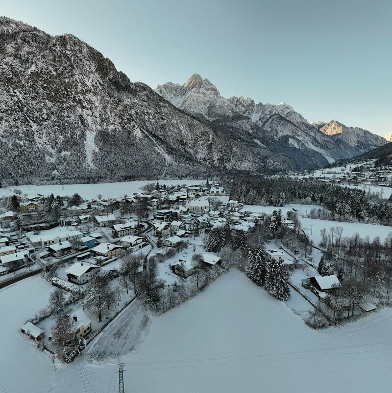 Ortsansicht Amlach Winter Das Bild zeigt die verschneite Ortschaft Amlach mit Blickrichtung ins Pustertal und auf die umliegende Bergwelt Osttirols. Die Häuser und Dächer der Siedlung sind dicht mit Schnee bedeckt. Rund um das Dorf erstrecken sich weitläufige, schneebedeckte Felder, Wiesen und Baumgruppen. Im Hintergrund ragen steile, felsige Gebirgskämme und schneebedeckte Gipfel der Lienzer Dolomiten empor.