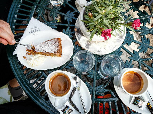 Aufnahme eines Tisches von oben, gedeckt mit 2 Tassen Kaffee, 2 Gläsern Wasser, einem Stück Kuchen mit Sahne und einer Blume im Topf.