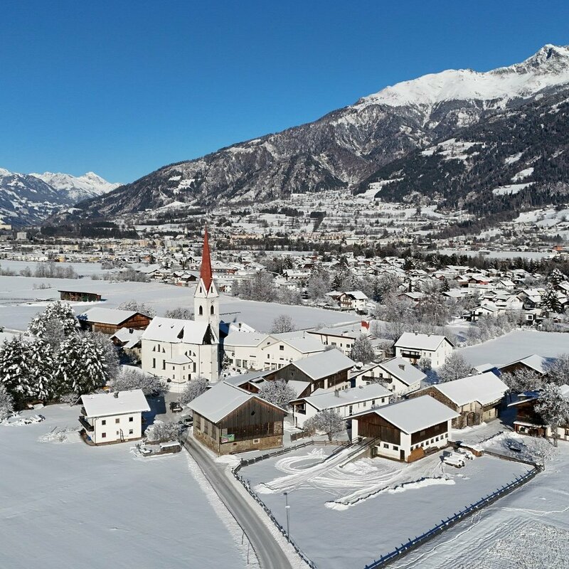 The photo shows the wintry landscape of the village of Tristach, looking toward Lienz. Traditional houses, farms, and residential buildings are clustered around the church, their roofs covered with an even layer of snow. The sky is brilliantly clear. 