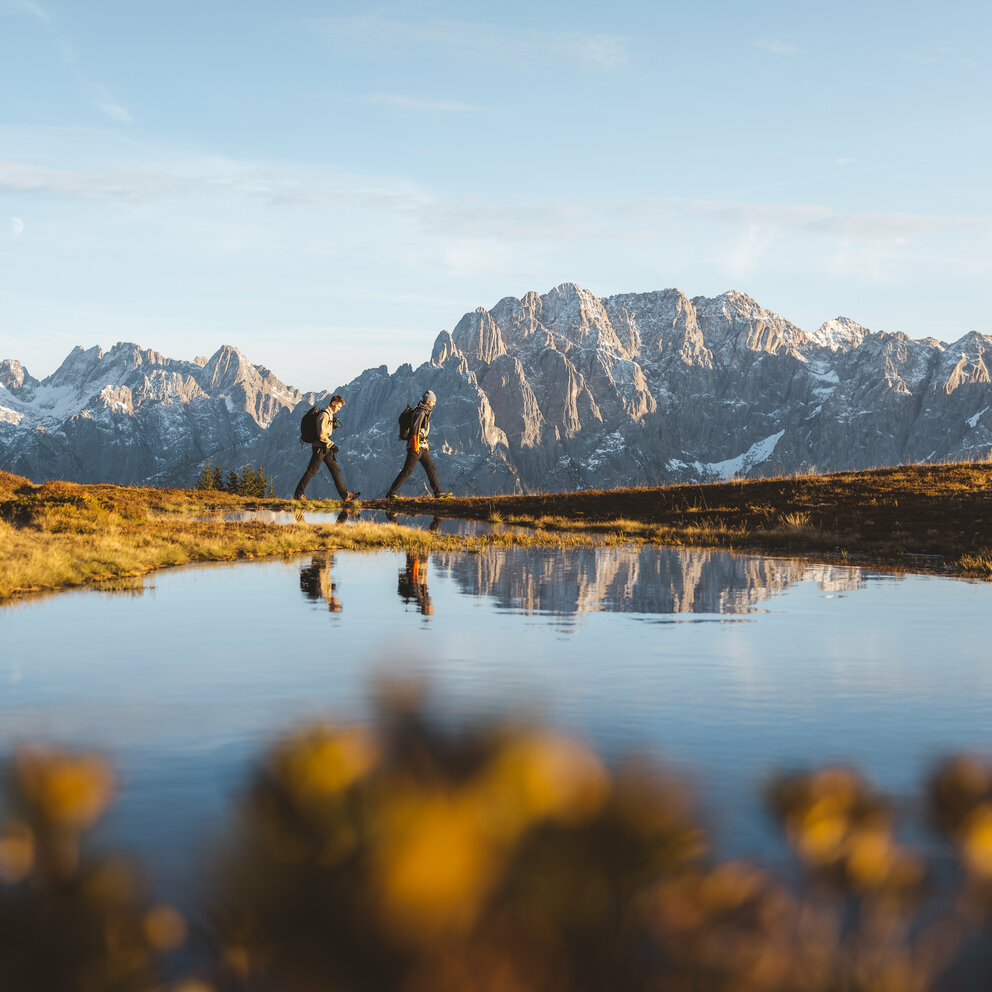 Zwei Personen spazieren am Bergsee am Hochstein bei traumhaftem Wetter mit Bergkulisse.