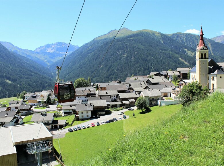 Das Dorf Obertilliach im Tiroler Gailtal, rechts im Bild die markante Kirche, links die bergwärts fahrende Gondelbahn vor alpiner Kulisse