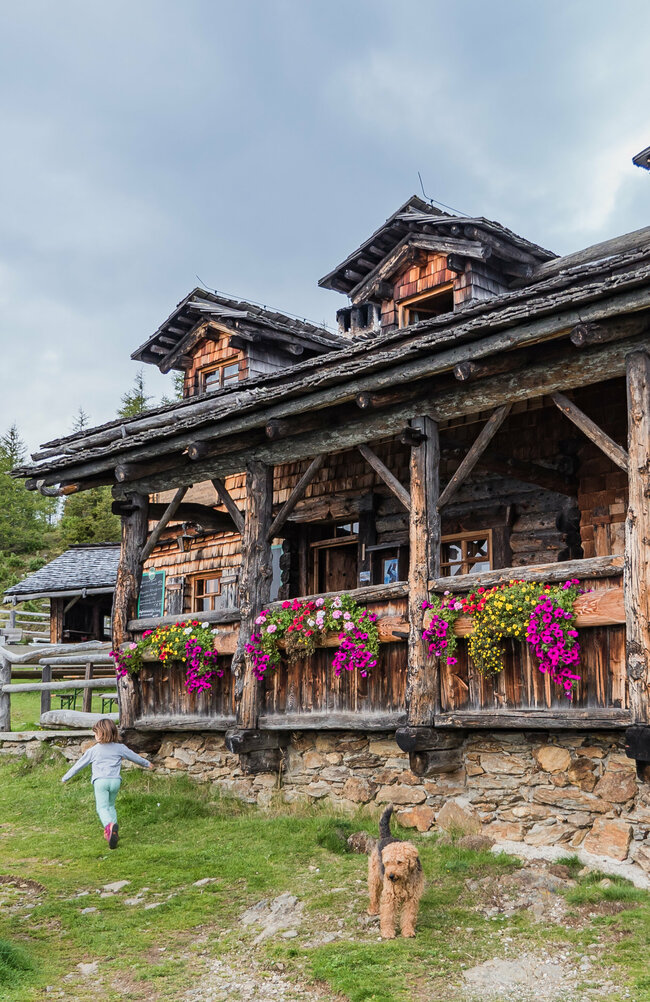 Anna Schutzhaus Das urige Anna Schutzhaus aus Holz mit Blumendeko und spielenden Kindern und einem Hund davor.