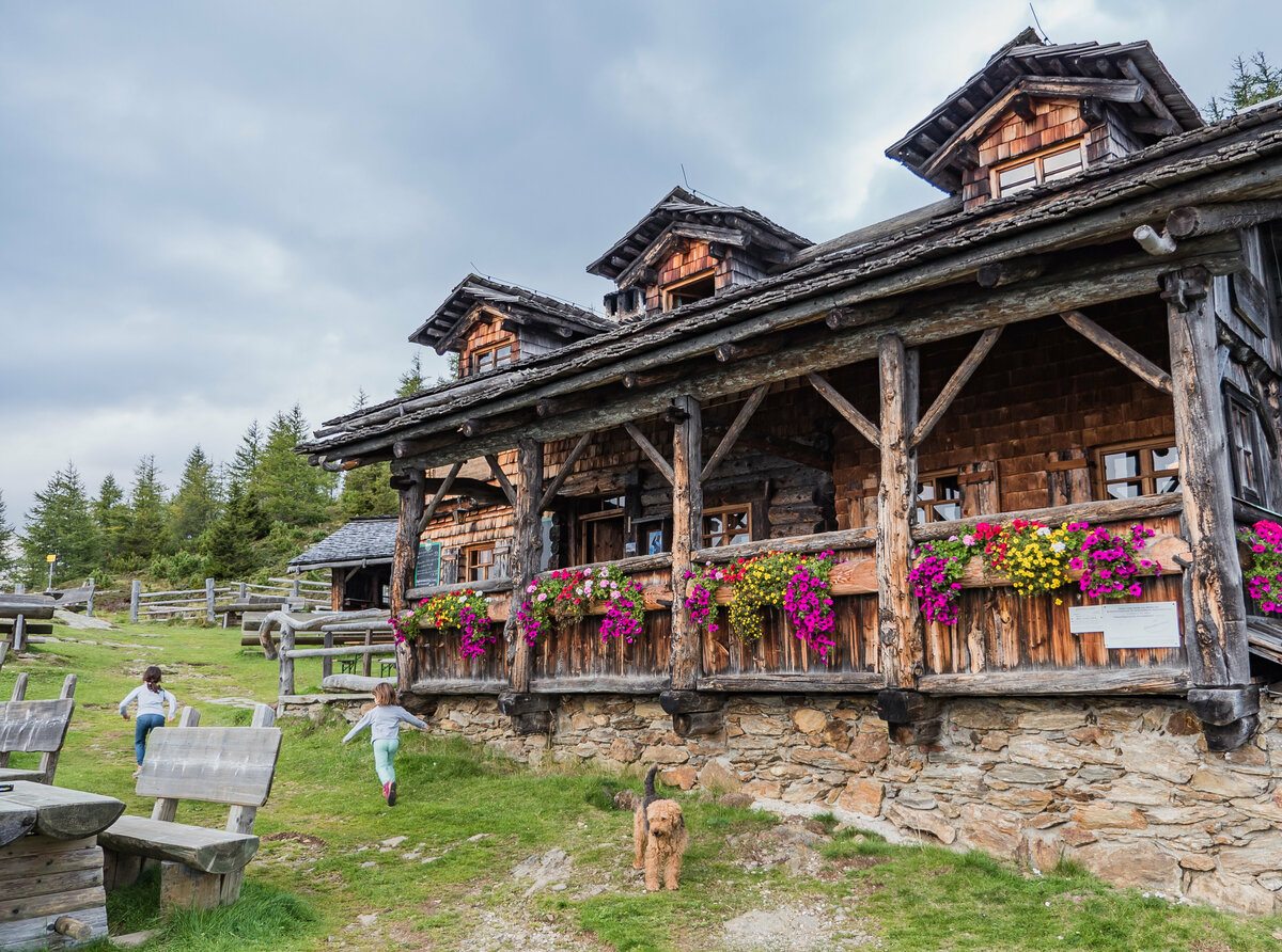 Das urige Anna Schutzhaus aus Holz mit Blumendeko und spielenden Kindern und einem Hund davor.