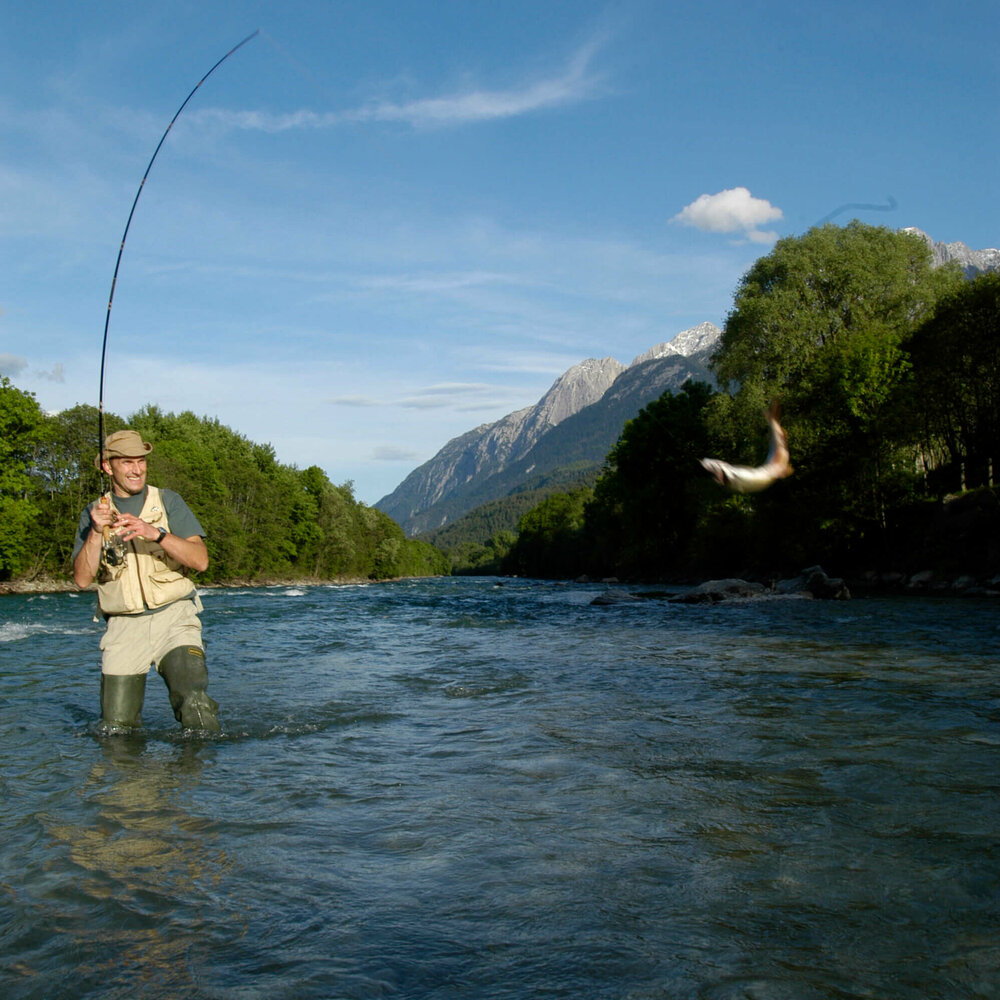 Ein Mann mit Hut steht in einem Fluss und ist am Fliegen fischen. An seiner Angel schwingt ein Fisch.