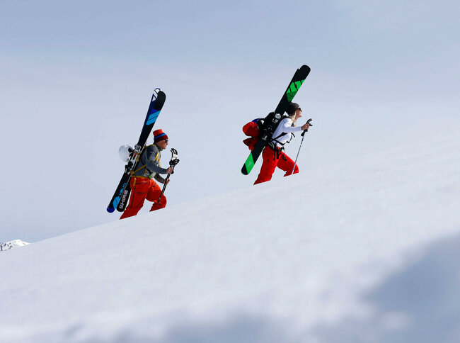Freeriden Ein Pärchen steigt mit Ski am Rücken einen schneebedeckten Berg hinauf. Beide tragen Mützen und rote Hosen. Die Ski sind schwarz-grün und schwarz-blau und sind an einem Rucksack befestigt.