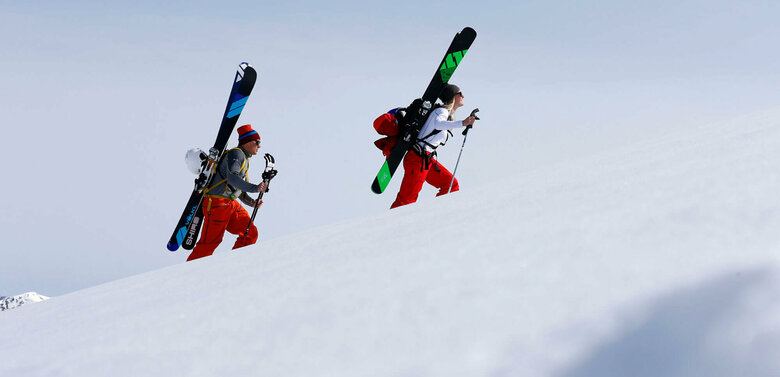 Ein Pärchen steigt mit Ski am Rücken einen schneebedeckten Berg hinauf. Beide tragen Mützen und rote Hosen. Die Ski sind schwarz-grün und schwarz-blau und sind an einem Rucksack befestigt.