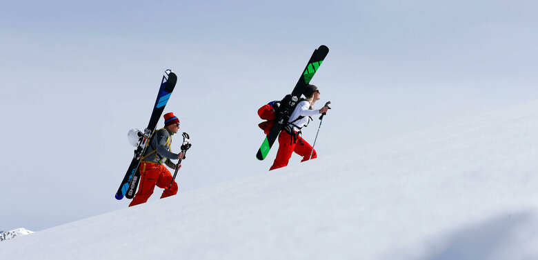 Ein Pärchen steigt mit Ski am Rücken einen schneebedeckten Berg hinauf. Beide tragen Mützen und rote Hosen. Die Ski sind schwarz-grün und schwarz-blau und sind an einem Rucksack befestigt.