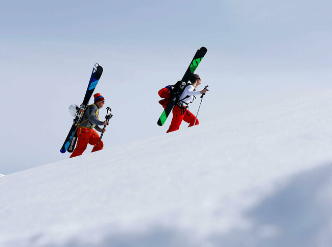 Freeriden Ein Pärchen steigt mit Ski am Rücken einen schneebedeckten Berg hinauf. Beide tragen Mützen und rote Hosen. Die Ski sind schwarz-grün und schwarz-blau und sind an einem Rucksack befestigt.