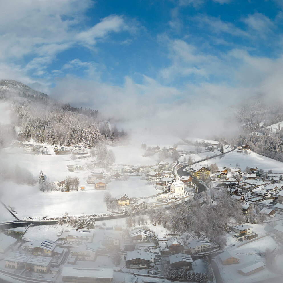 Luftaufnahme von der Ortschaft Iselsberg im Winter. Einige Wolken ziehen am Himmel vorbei.