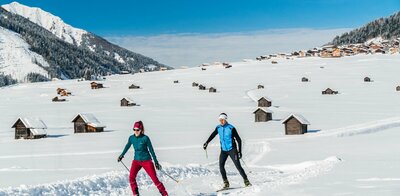 Zwei Wintersportler laufen auf ihren Langlaufskiern auf der Loipe in den Obertilliacher Feldern mit den zahlreichen kleinen Hütten und dem Dorf im Hintergrund