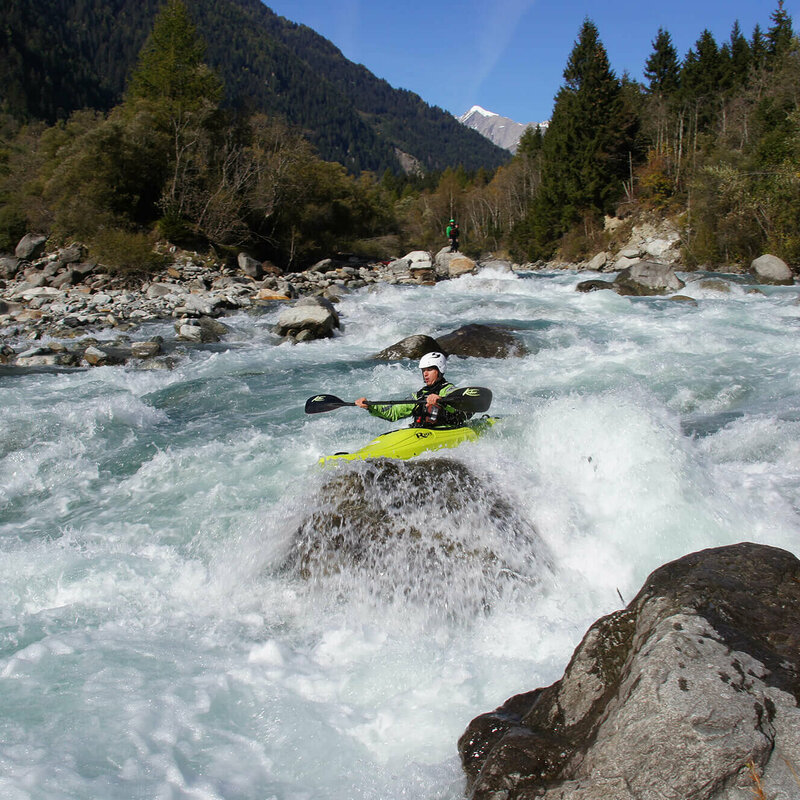 Ein Kajakfahrer inmitten des Wildwasserflusses Isel in Osttirol, dahinter ist Wald und eine Bergspitze.