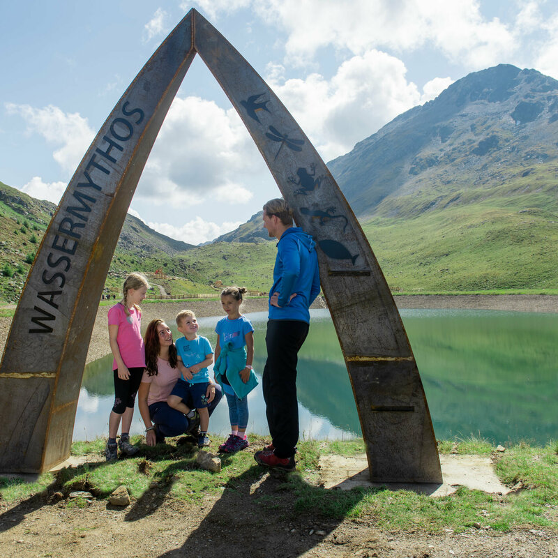Eine Familie beim Wassermythos Ochsenlacke im Skizentrum St. Jakob i. D.. Sie stehen an dem kunstvollen Bogen welcher am Rande des Ufers errichtet wurde. Die Sonne lässt die umliegende Bergwelt und das Wasser in einem warmen Licht erstrahlen.