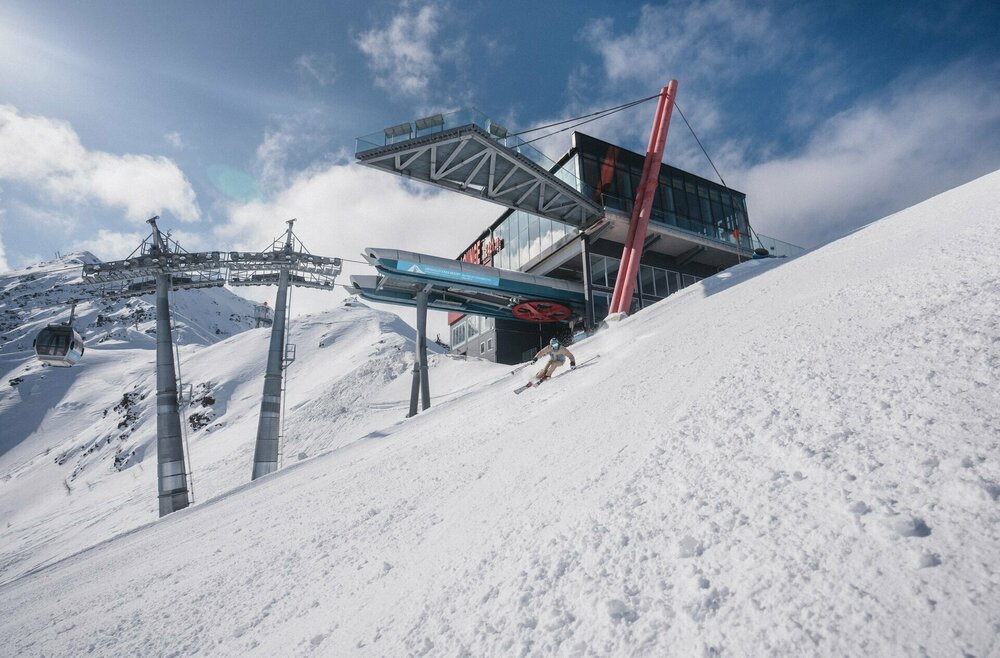Skifahren GG Resort Kals-Matrei Ein Skifahrer fährt eine Piste hinunter, im Hintergrund sieht man eine Gondelbahn und eine Berghütte.