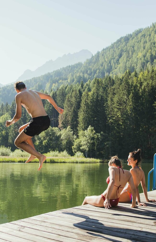 Zwei Mädchen sitzen auf einem Holzsteg und ein Junge springt ins Wasser am Tristacher See.